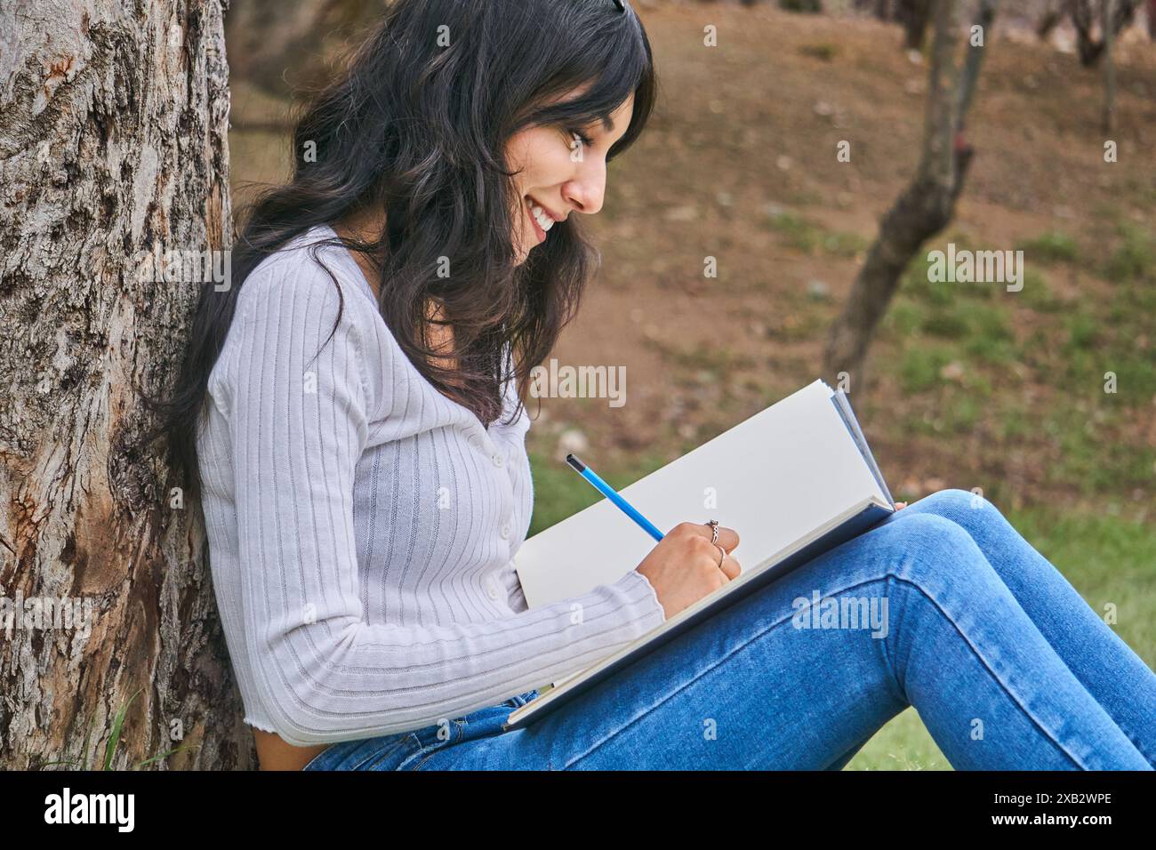 Une femme contente aime dessiner dans un parc serein, assise contre un arbre Banque D'Images
