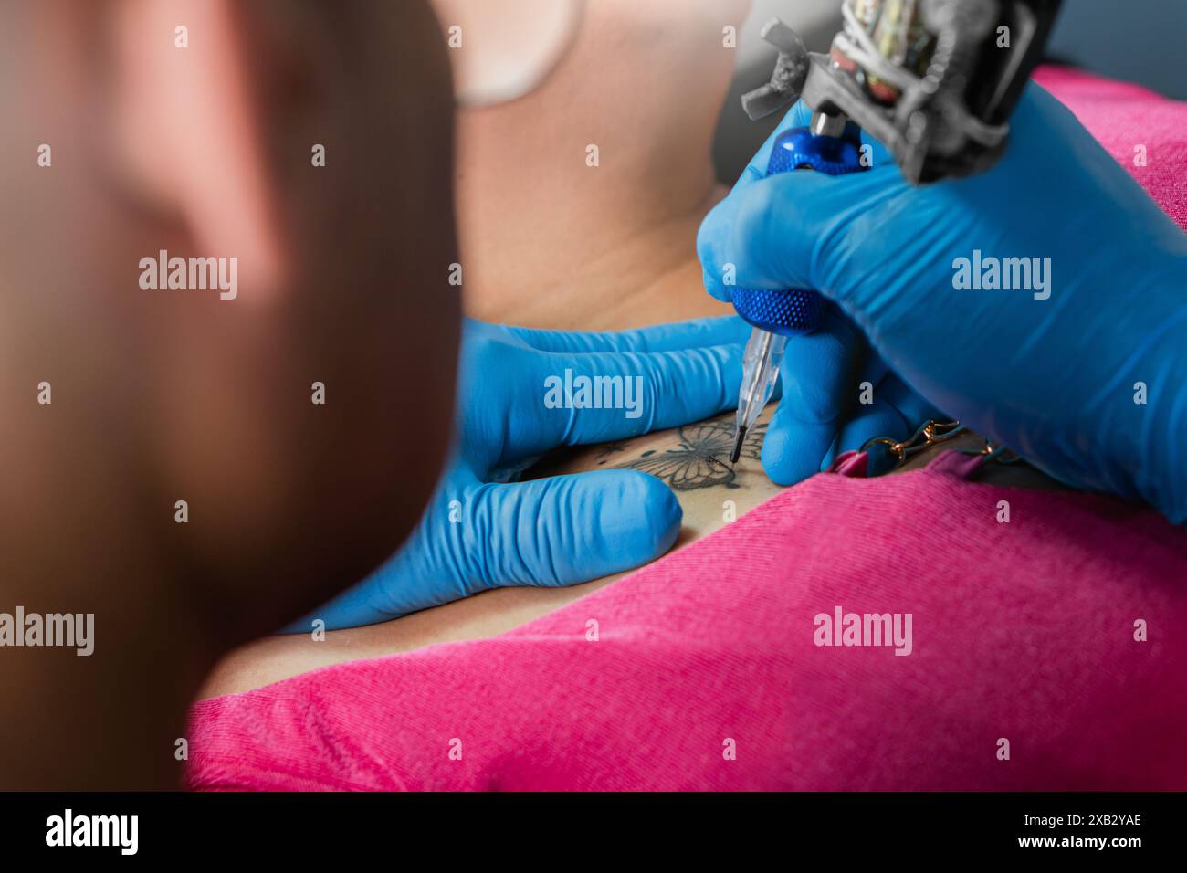 Image recadrée méconnaissable d'un tatoueur dans des gants bleus encrant un dessin sur l'épaule d'un client, démontrant le processus complexe du tatouage Banque D'Images