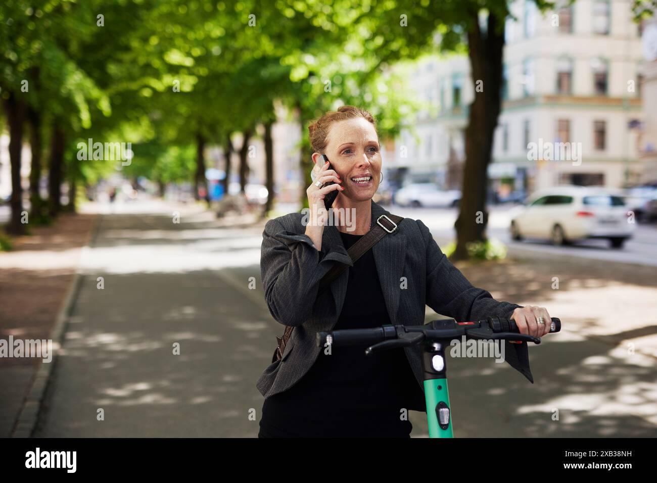 Femme d'affaires souriante avec scooter électrique poussant parler sur téléphone intelligent sur le sentier pédestre Banque D'Images