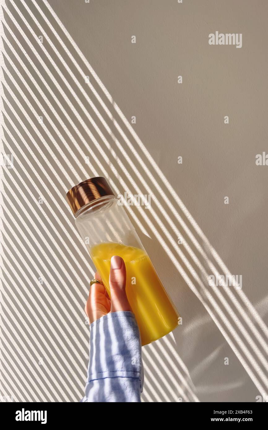 Main féminine tenant une bouteille réutilisable avec du jus d'orange sur le mur dans la lumière du matin et l'ombre des stores Banque D'Images