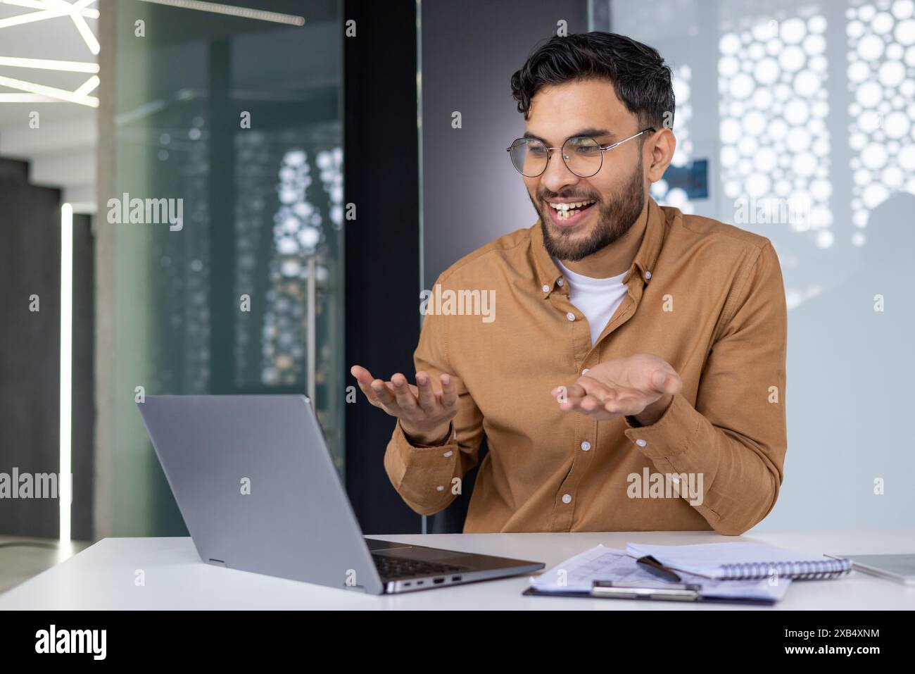 Souriant jeune homme d'affaires indien, employé de bureau assis à un bureau et communiquant à distance sur une caméra d'ordinateur portable, faisant des gestes avec ses mains, tenant une réunion en ligne. Banque D'Images