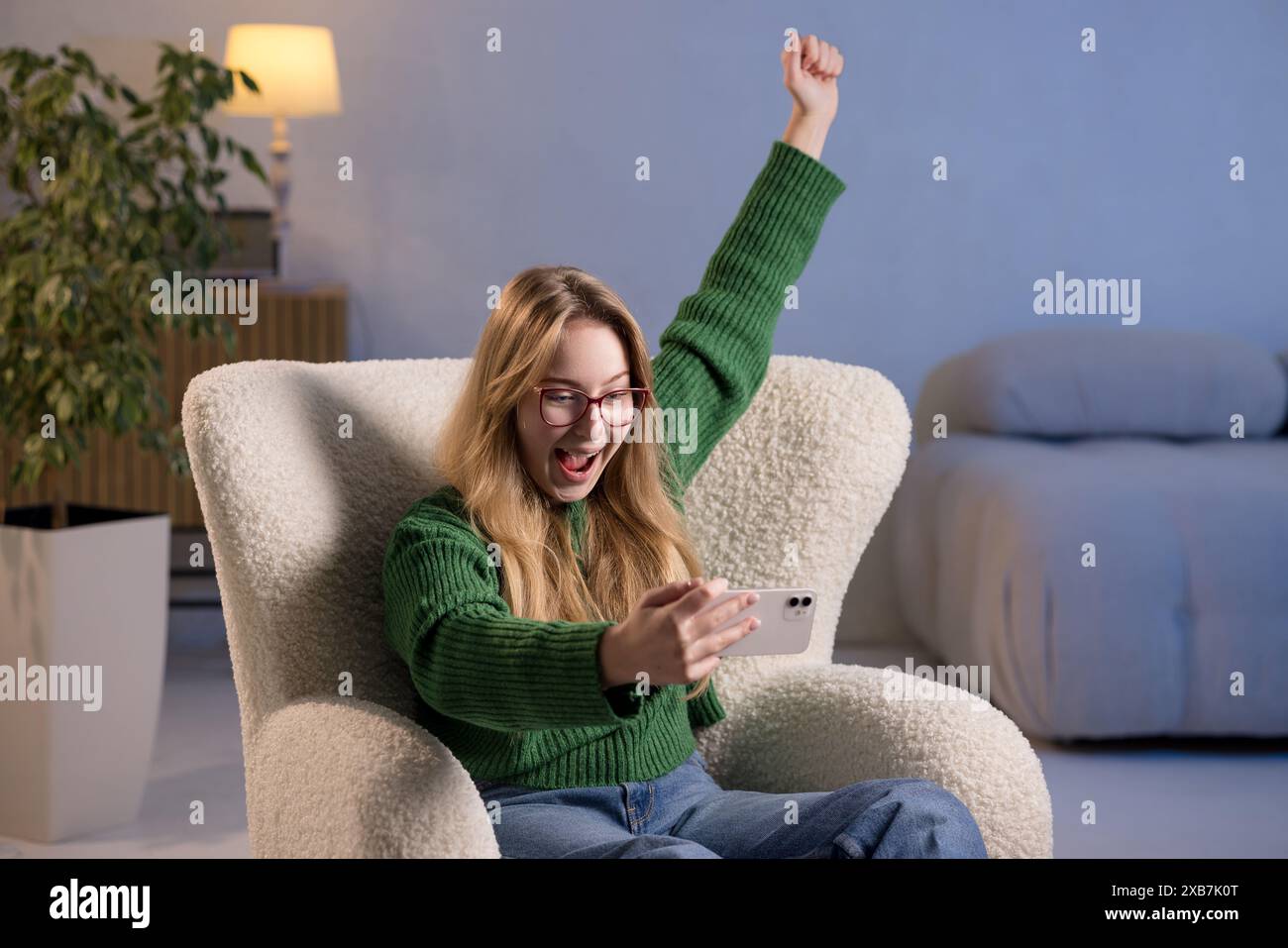 Une femme est confortablement assise sur une chaise, tenant un téléphone portable avec ses bras levés. La salle a un canapé, des plantes, et une atmosphère joyeuse Banque D'Images