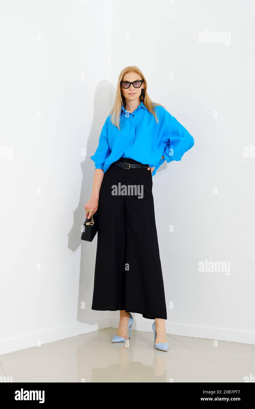 Femme d'affaires élégante en chemise boutonnée bleue et jupe noire ample debout à côté du mur blanc Banque D'Images