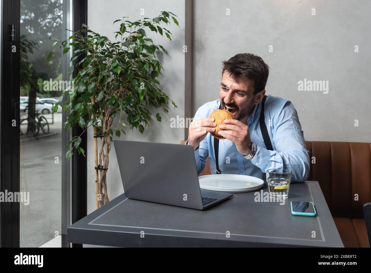 Jeune homme d'affaires affamé à la pause déjeuner assis à la cafétéria mangeant hamburger et utilisant un ordinateur portable pour le travail en ligne. Amoureux des hamburgers homme d'affaires manger et Banque D'Images