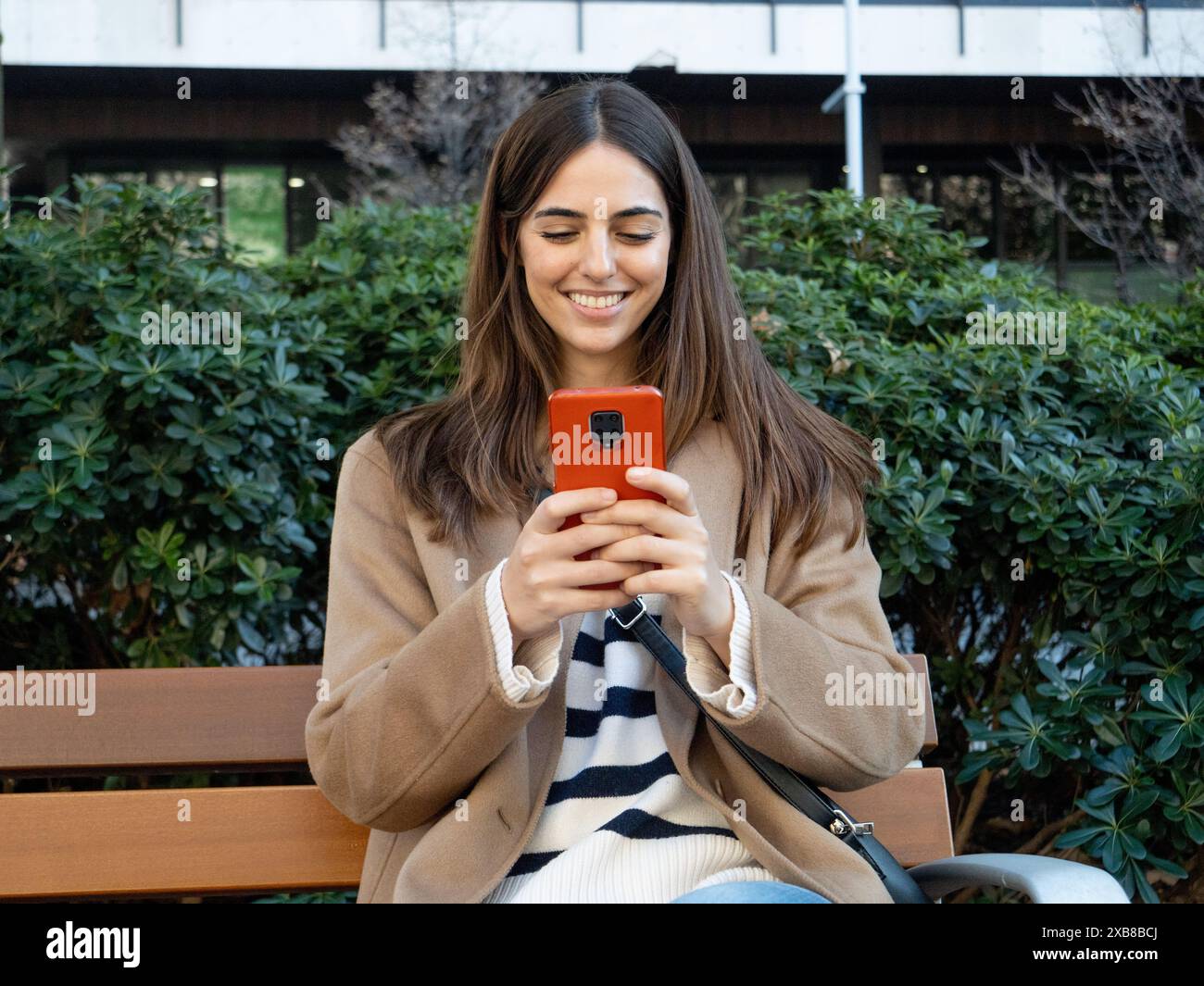Jeune femme élégante souriante assise sur un banc dans une ville discutant avec un smartphone Banque D'Images