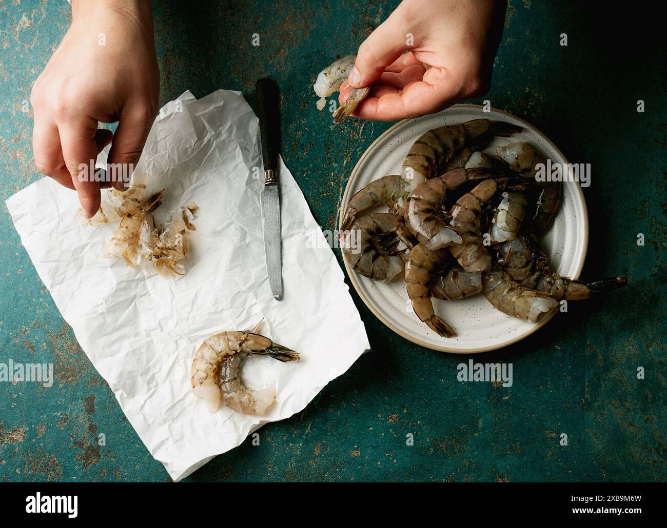 Mains épluchant des crevettes sur une assiette avec un couteau, et des coquilles de crevettes sur du papier froissé sur un fond sarcelle. La photo capture une étape dans la crevette prepa Banque D'Images