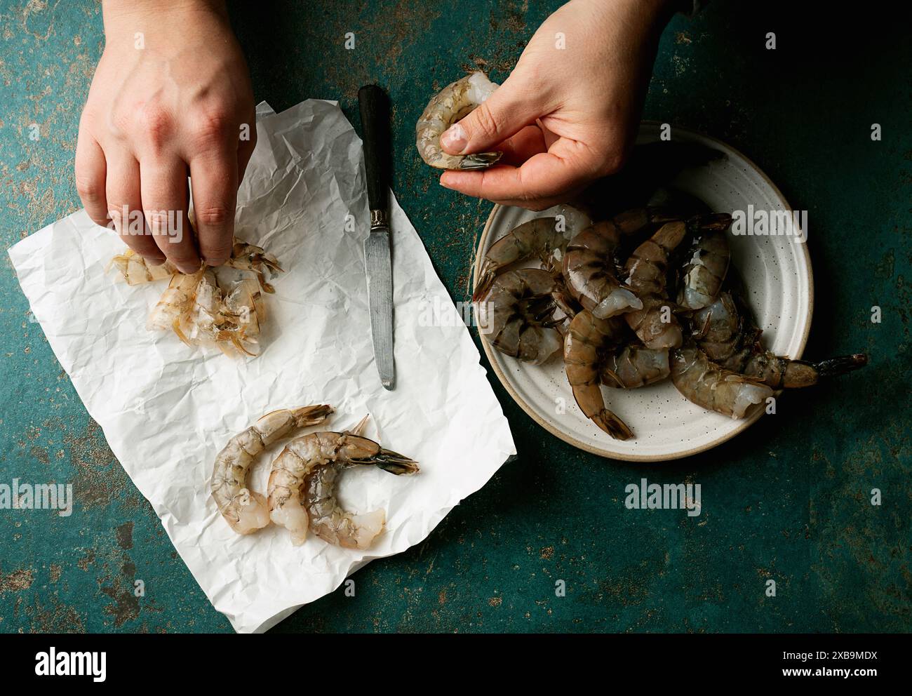 Mains épluchant des crevettes sur une assiette avec un couteau, et des coquilles de crevettes sur du papier froissé sur un fond sarcelle. La photo capture une étape dans la crevette prepa Banque D'Images