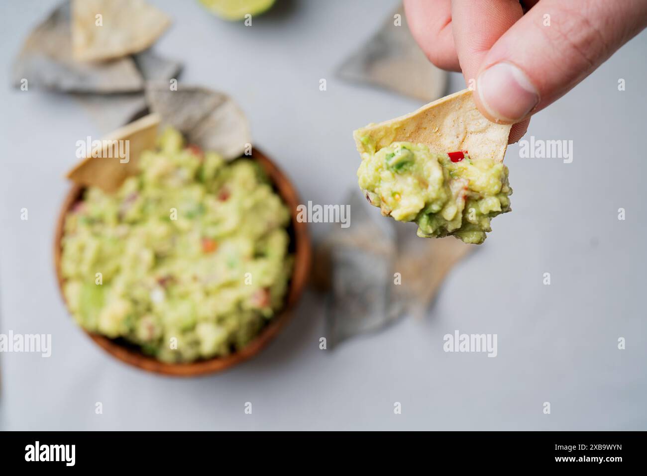 Main tenant une puce de tortilla au guacamole au-dessus d'un bol en bois rempli de guacamole, garni de coriandre. Coin de citron vert et chips tortilla de maïs bleu Banque D'Images
