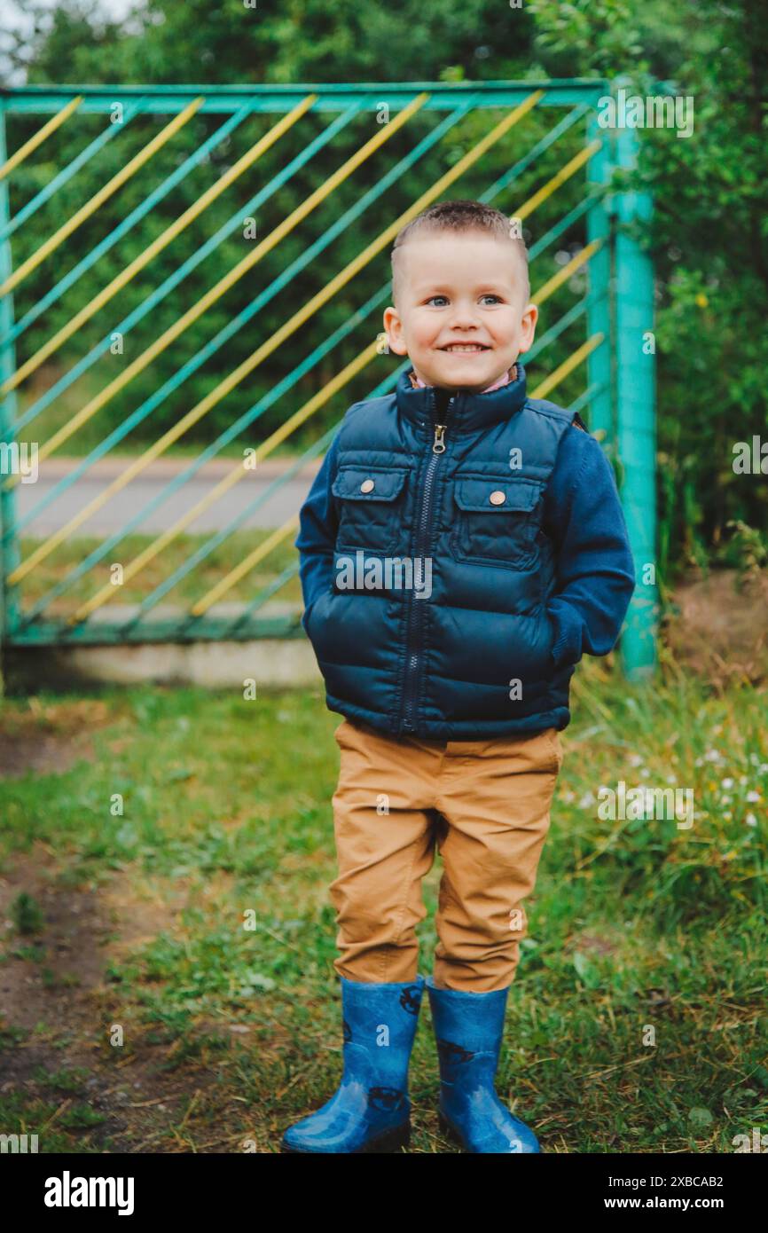 Jeune garçon portant une veste bleue et des bottes se tient à l'extérieur près d'un gate vert.Belarus.Minsk Banque D'Images