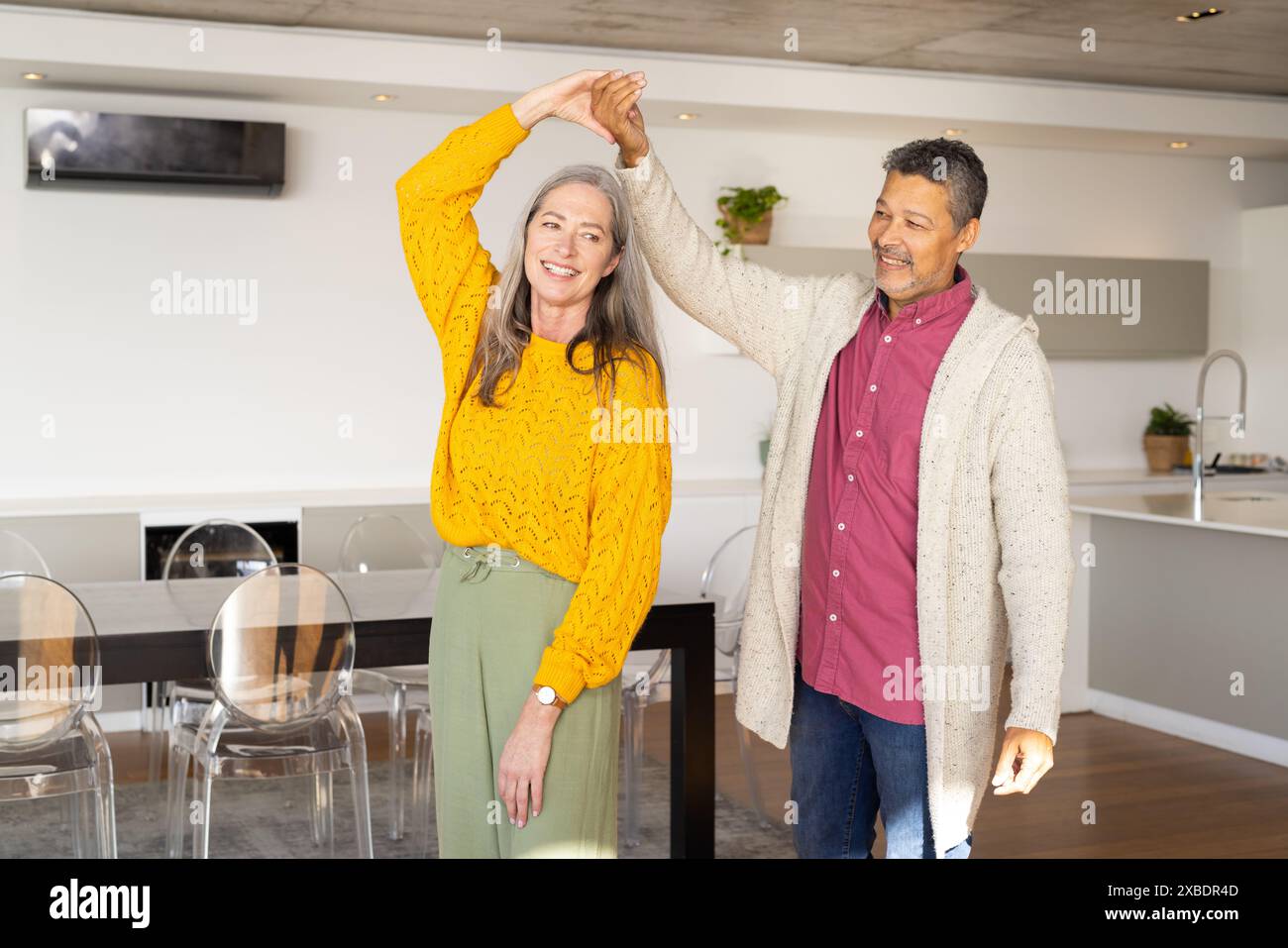 Un couple biracial mature dansant ensemble, souriant chaleureusement l'un à l'autre, à la maison. Ils sont dans une cuisine moderne et lumineuse avec des chaises claires et un grand din Banque D'Images