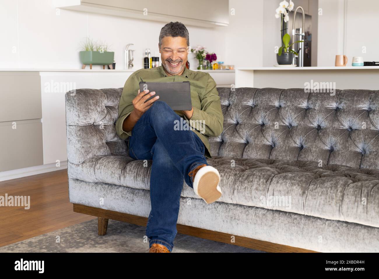 Homme biracial mature assis sur le canapé, souriant tout en utilisant une tablette, à la maison. Salon moderne avec une cuisine en arrière-plan, avec des plantes et sty Banque D'Images