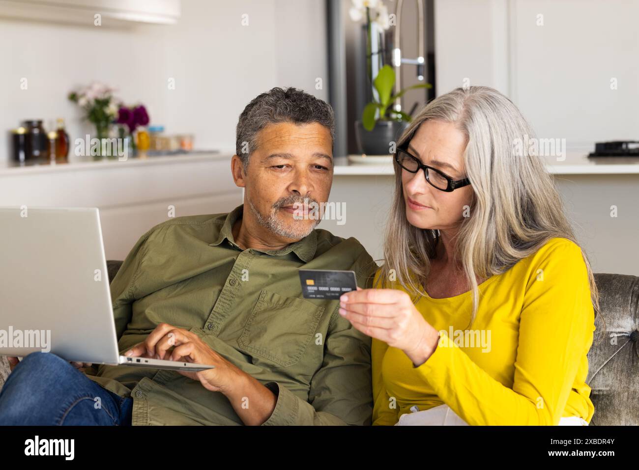 Un couple mature biracial utilise un ordinateur portable et une carte de crédit ensemble dans une cuisine moderne, à la maison Banque D'Images