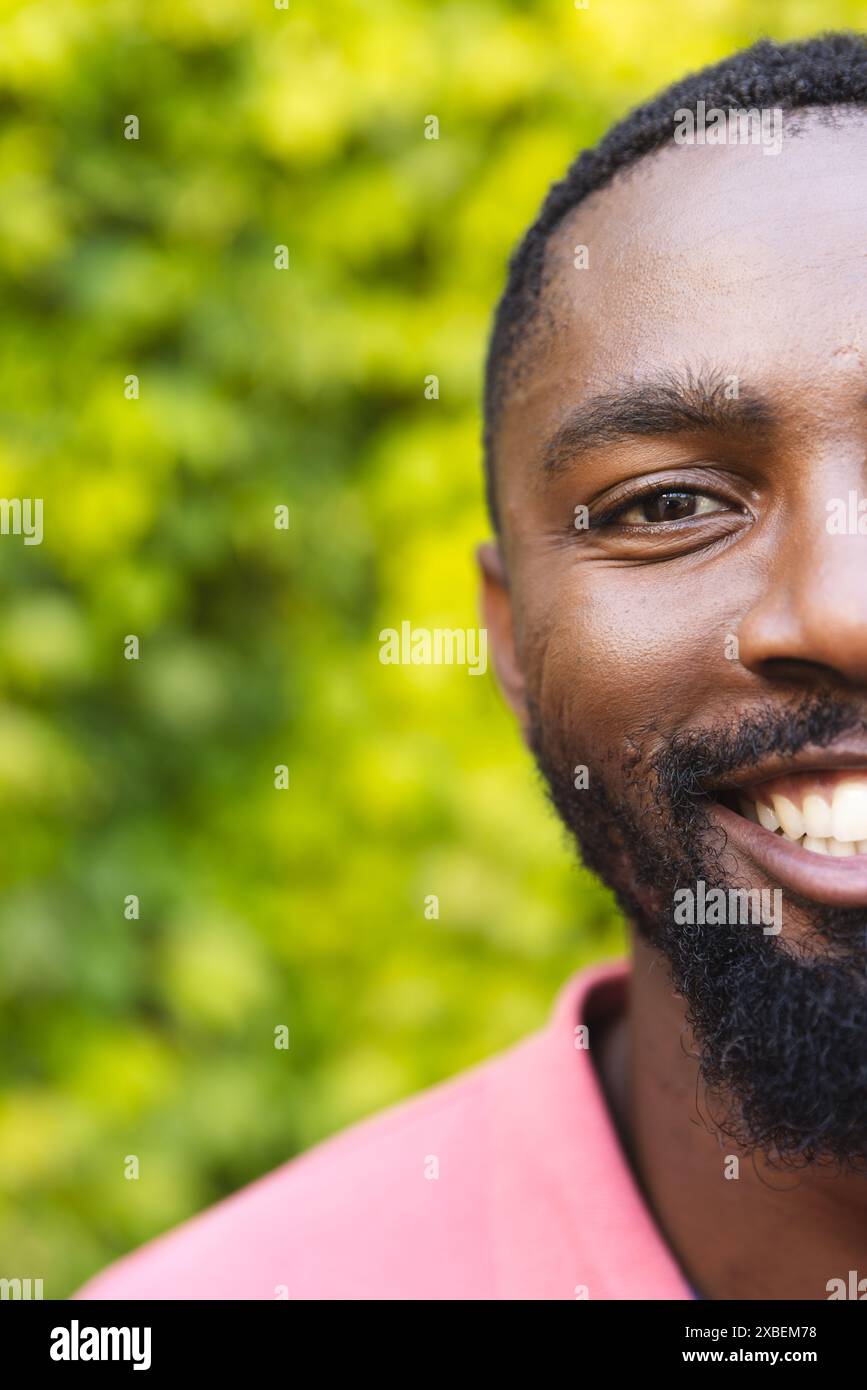 Jeune homme afro-américain souriant chaleureusement, portant une chemise rose, à la maison. Feuillage vert luxuriant créant un fond vert naturel et vibrant, ajoutant à liv Banque D'Images Jeune homme afro-américain souriant chaleureusement, portant une chemise rose, à la maison. Feuillage vert luxuriant créant un fond vert naturel et vibrant, ajoutant à liv Banque D'Images