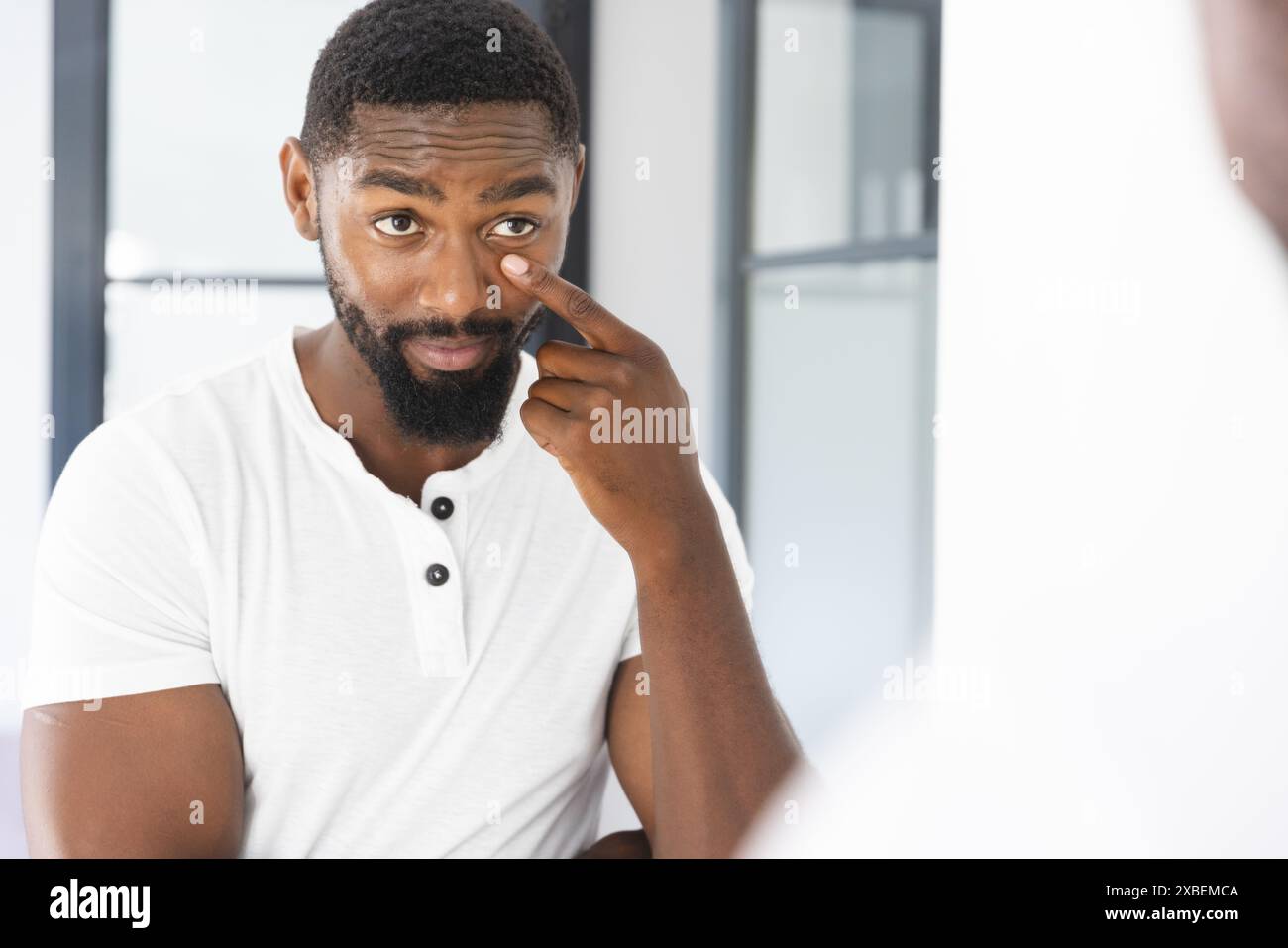Jeune homme afro-américain avec la barbe touchant son visage tout en se regardant dans un miroir, à la maison Banque D'Images