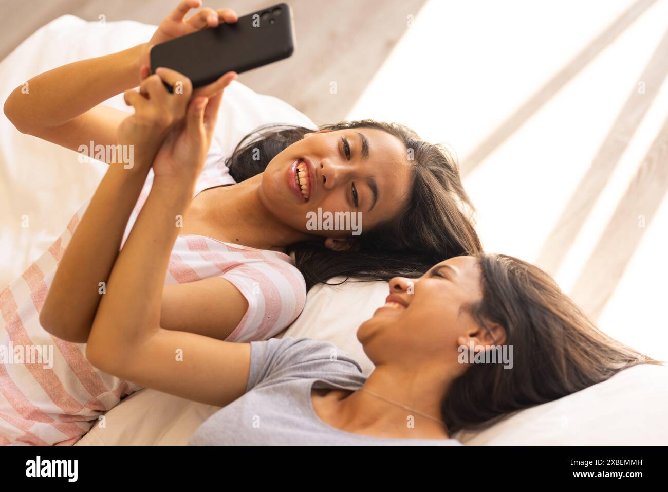 Deux jeunes sœurs biraciales sont allongées sur un lit, souriantes et partageant un moment avec un téléphone, à la maison Banque D'Images