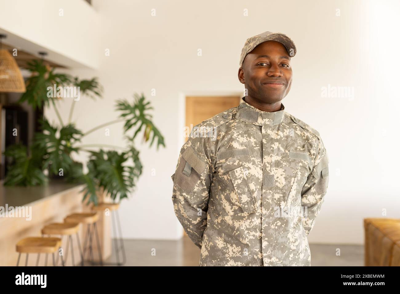 Jeune soldat afro-américain en uniforme militaire debout à l'intérieur, souriant avec confiance. Intérieur moderne avec meubles en bois et grand p vert Banque D'Images Jeune soldat afro-américain en uniforme militaire debout à l'intérieur, souriant avec confiance. Intérieur moderne avec meubles en bois et grand p vert Banque D'Images