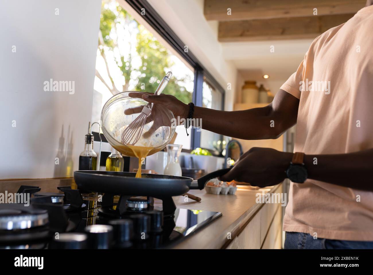 Homme afro-américain cuisinant des œufs pour le petit déjeuner dans la cuisine moderne. de grandes fenêtres offrent une vue sur un jardin ensoleillé, créant une atmosphère lumineuse et aérée Banque D'Images