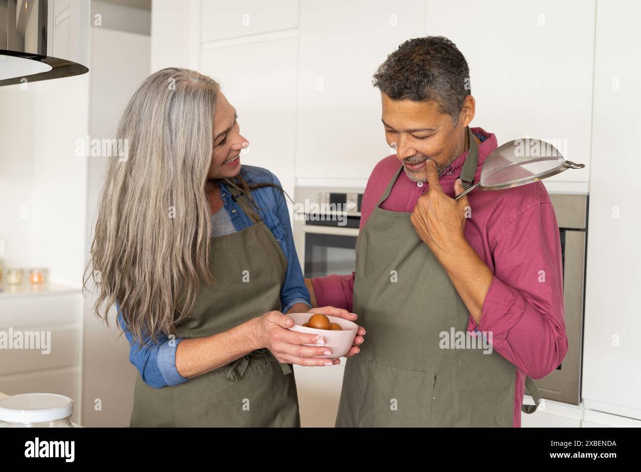 Un couple biracial mature cuit ensemble dans leur cuisine, à la maison. porter des tabliers assortis, debout dans une cuisine moderne avec des armoires blanches et sta Banque D'Images