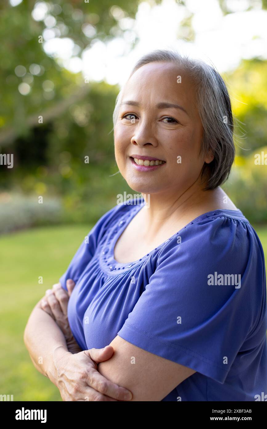 Femme aînée asiatique souriante avec les bras croisés, apparaissant satisfaite et détendue, à la maison Banque D'Images