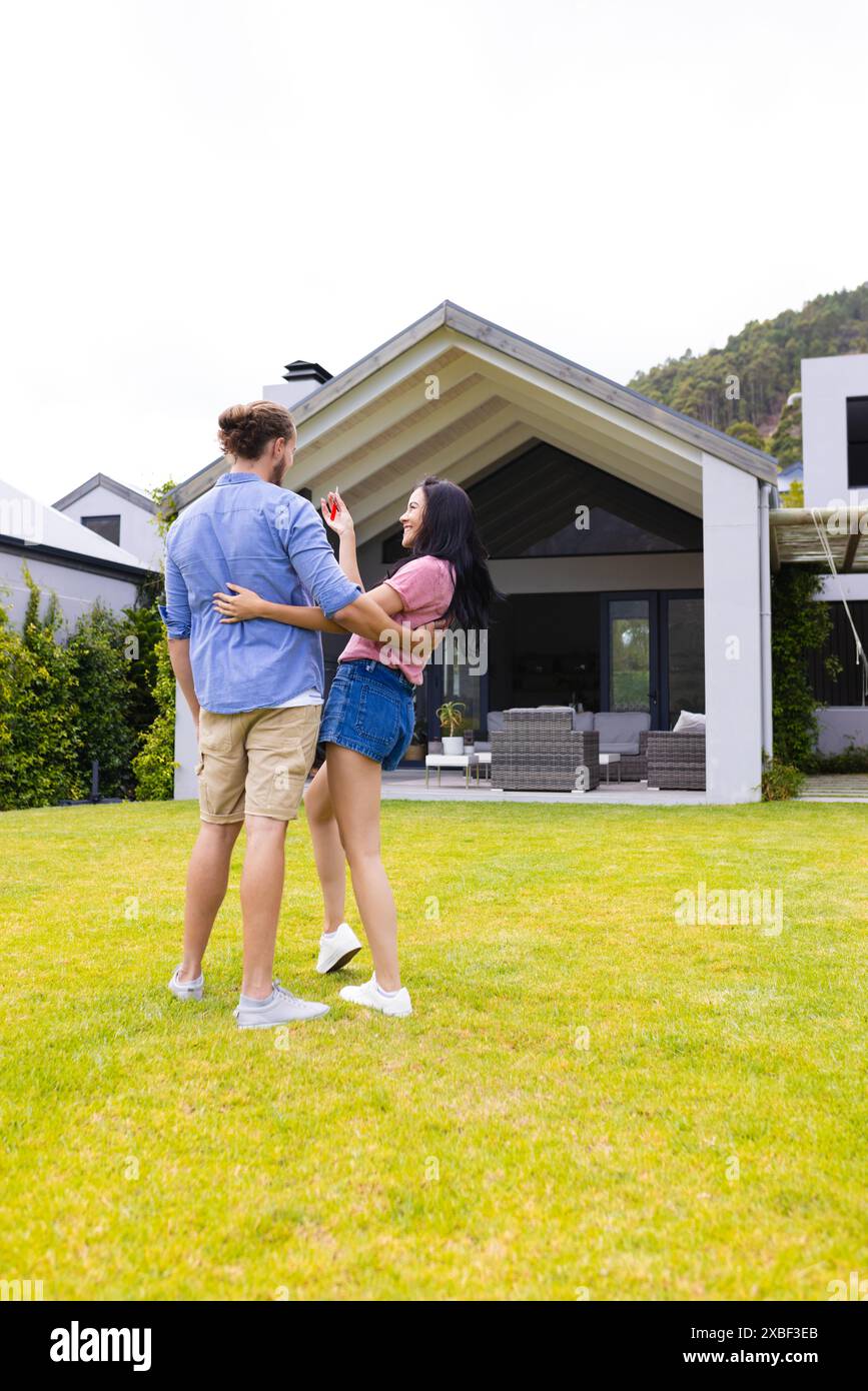 Jeune couple diversifié embrasser devant une maison moderne, achetant la première maison. Ils sont debout sur une pelouse verte luxuriante avec un mobilier d'extérieur contemporain Banque D'Images Jeune couple diversifié embrasser devant une maison moderne, achetant la première maison. Ils sont debout sur une pelouse verte luxuriante avec un mobilier d'extérieur contemporain Banque D'Images