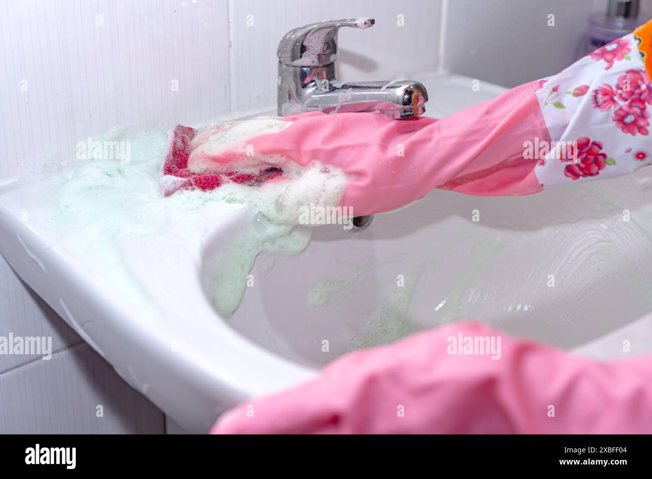 Femme dans des gants roses nettoyant évier en céramique blanche avec éponge et détergent, gros plan Banque D'Images