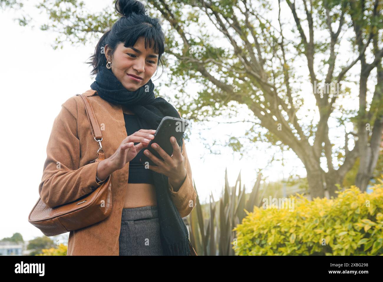 Une femme latine heureuse des notifications sur son téléphone portable. Elle porte une veste marron et une écharpe noire, défilant et tapant sur l'écran pendant Banque D'Images