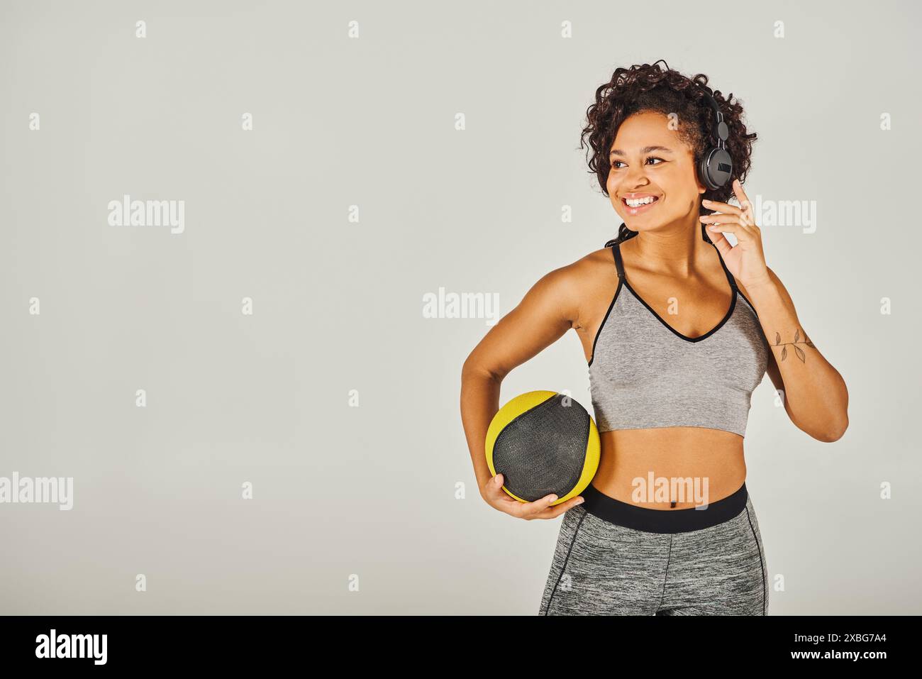 Femme afro-américaine aux cheveux bouclés en vêtements de sport, portant des écouteurs, tenant une balle, Banque D'Images