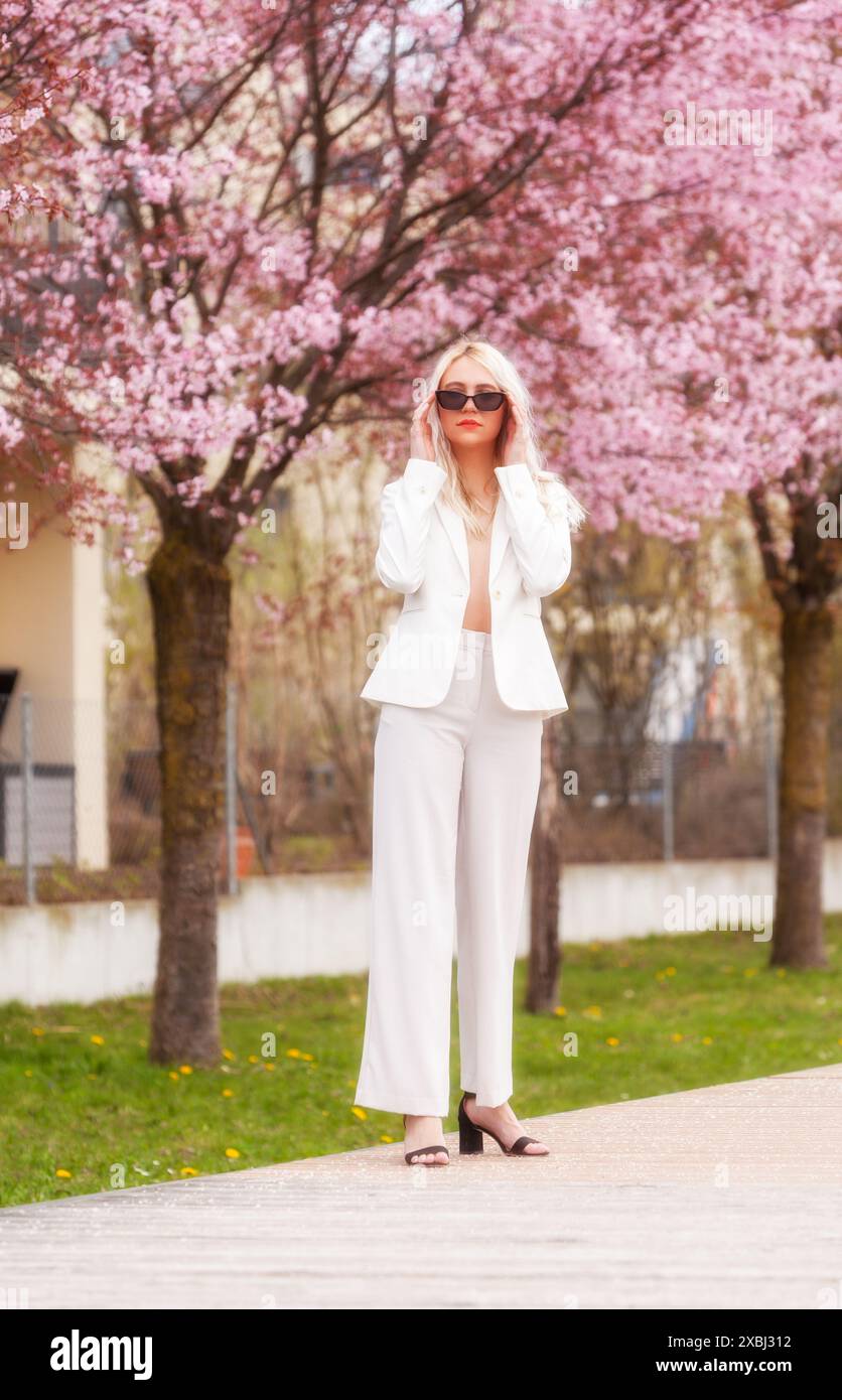 Une femme élégante dans un costume de pantalon blanc se tient devant un arbre en fleurs et met ses lunettes de soleil. La scène combine la beauté naturelle avec la mode Banque D'Images