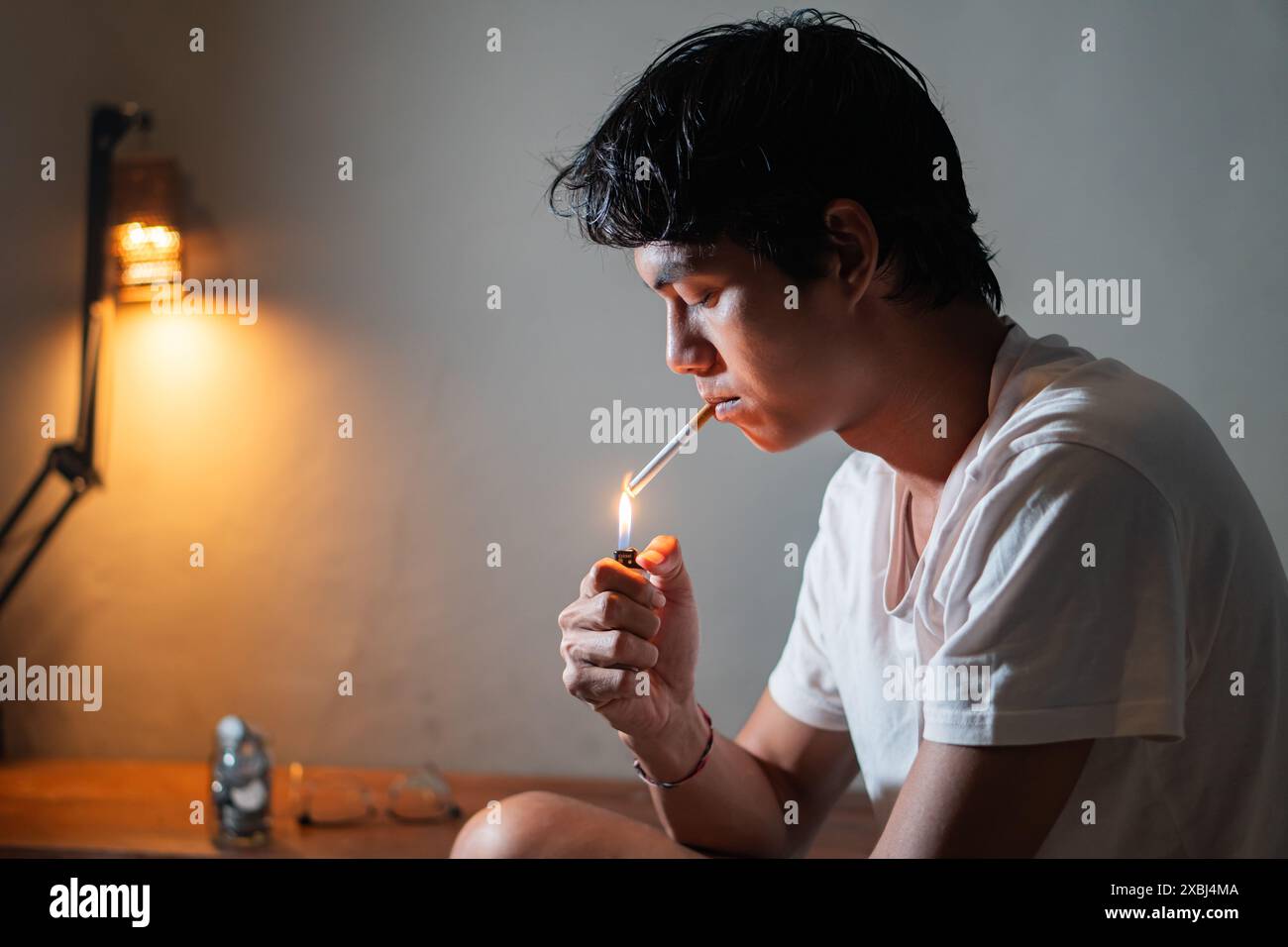 Un jeune homme allumant une cigarette avec un briquet dans une pièce faiblement éclairée. Banque D'Images