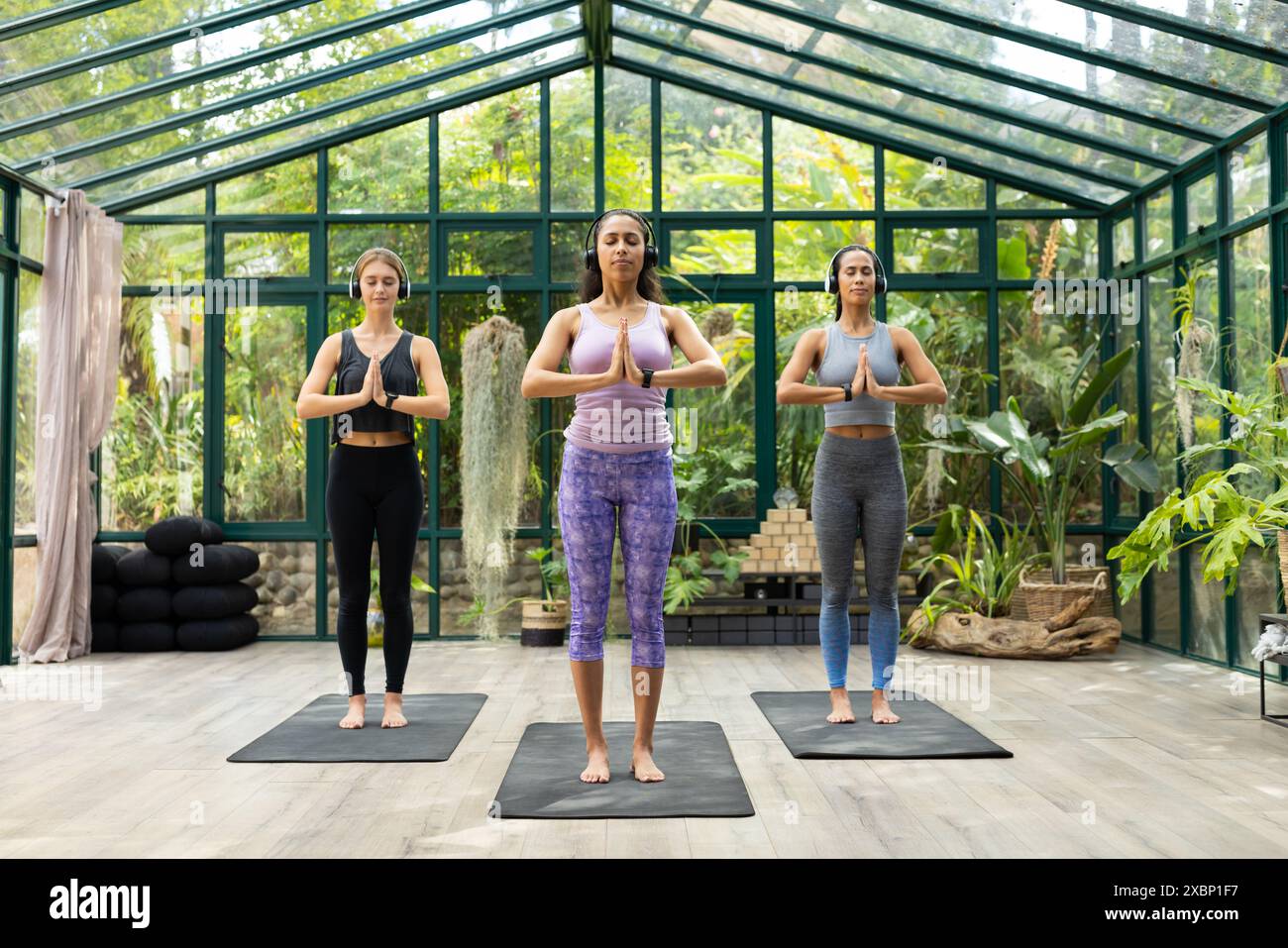 Trois femmes en forme pratiquant le yoga dans une maison de verre entourée de verdure luxuriante Banque D'Images