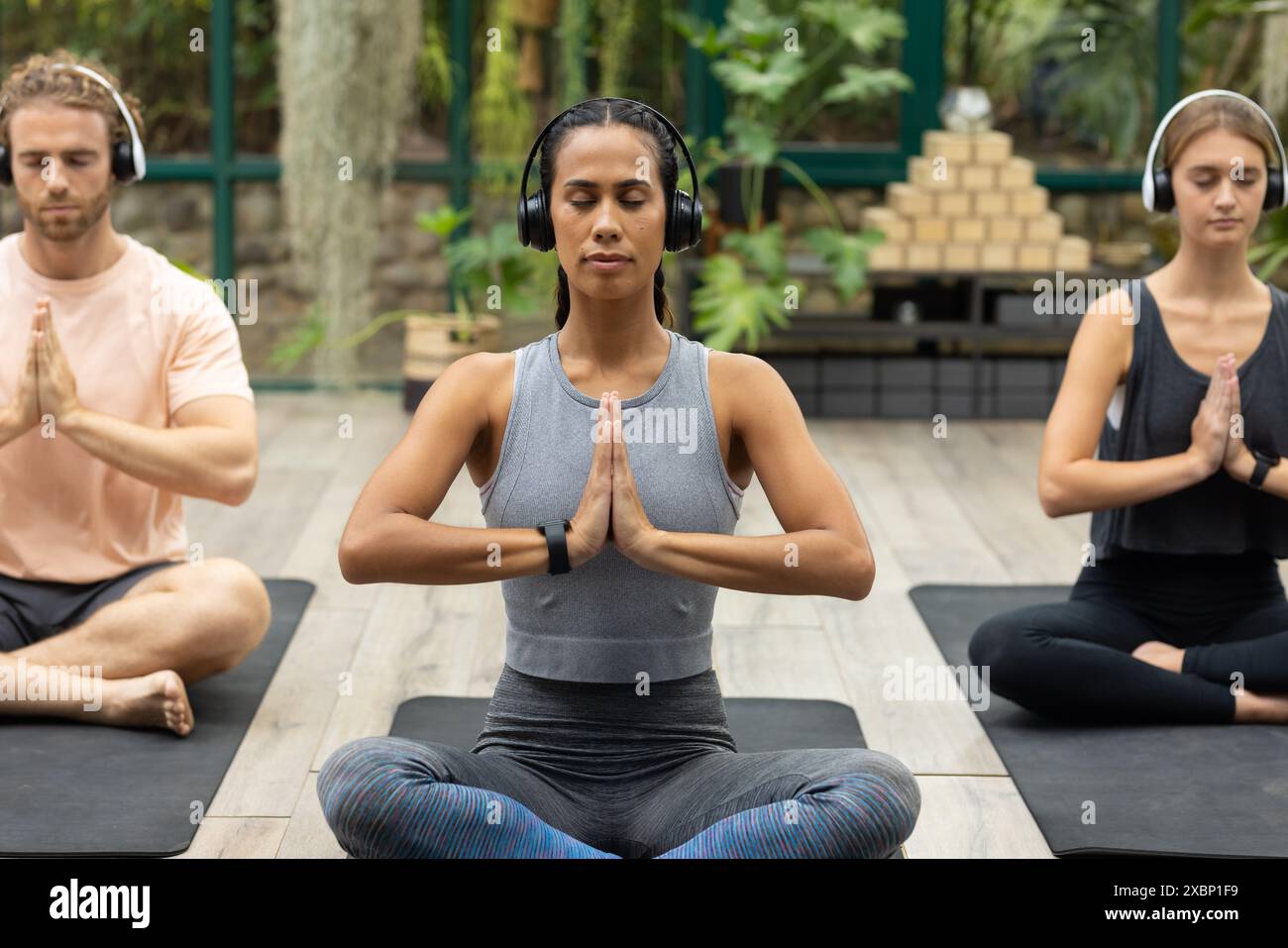 Diverses personnes au cours de yoga méditant avec des écouteurs dans une maison de verre sereine Banque D'Images