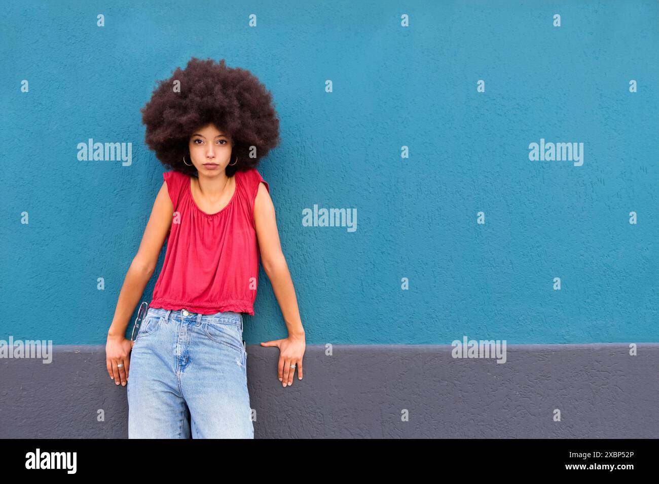 Jeune femme marocaine confiante avec des cheveux afro regardant la caméra tout en se tenant debout et appuyée avec les mains sur le mur sur fond bleu à la lumière du jour Banque D'Images