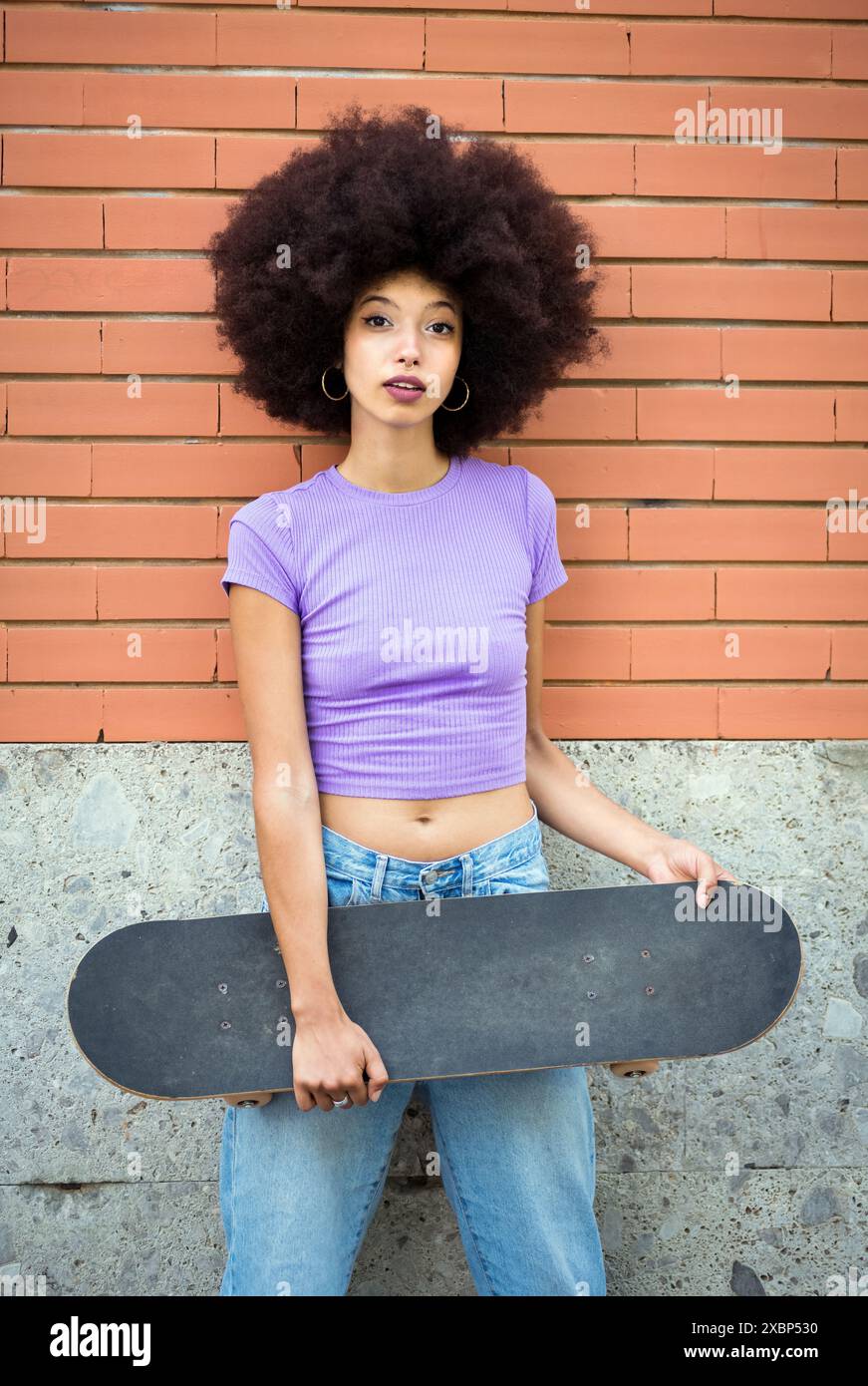 Confiante jeune femme afro-américaine avec coiffure afro dans des vêtements décontractés debout avec skateboard contre le mur de briques et regardant la caméra Banque D'Images