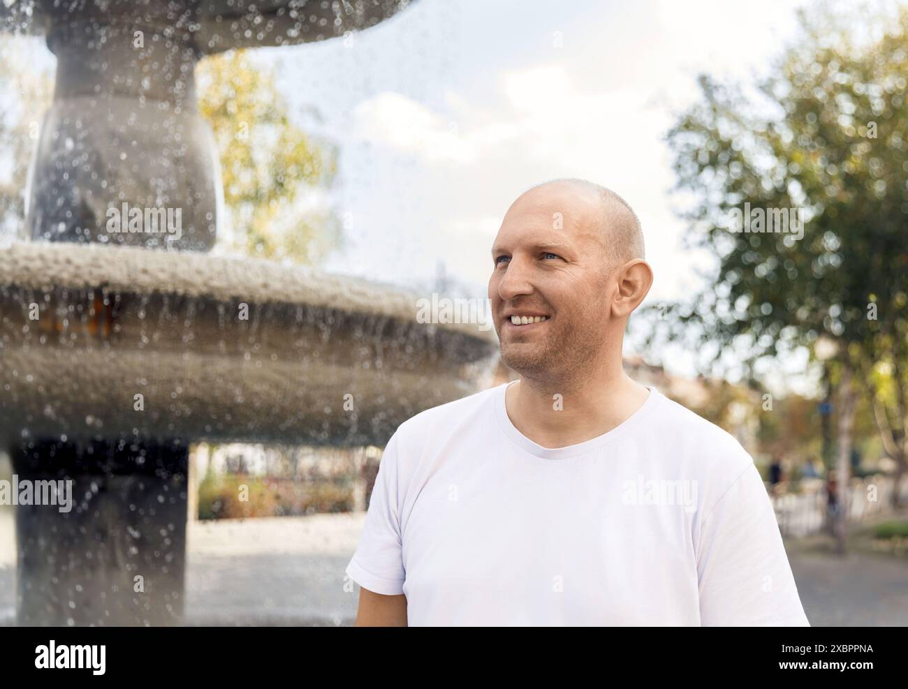 Homme chauve en t-shirt blanc souriant près de la fontaine, parc extérieur, journée ensoleillée Banque D'Images