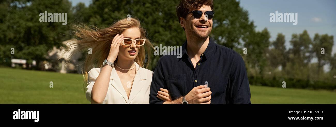 Un jeune homme et une femme glamour dans une tenue élégante marchent à travers un champ luxuriant, arborant des lunettes de soleil tendance sous le soleil. Banque D'Images