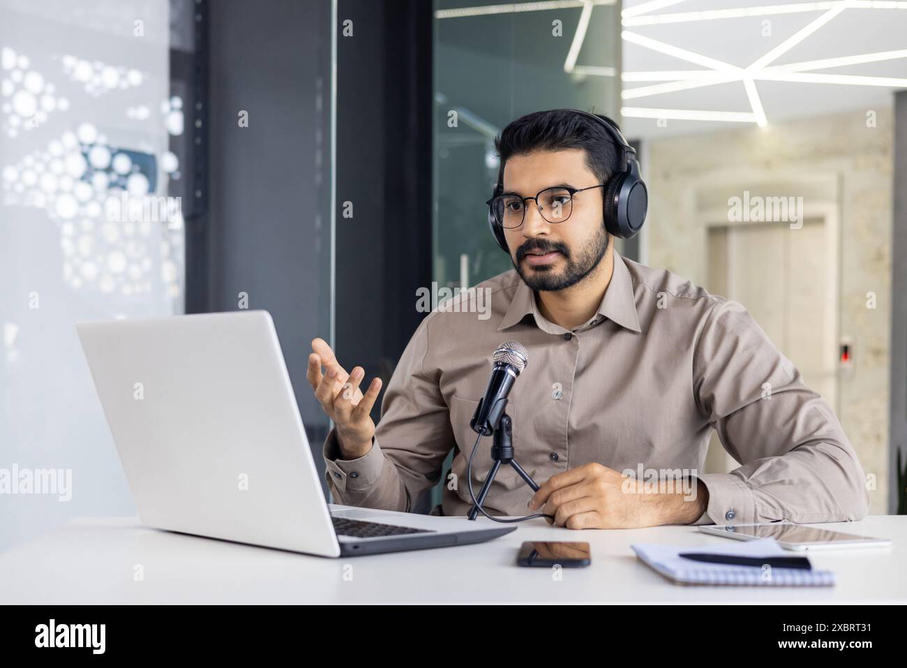 Jeune homme indien assis au bureau dans le bureau portant un casque et parlant dans le microphone lors d'un appel vidéo sur ordinateur portable, faisant des gestes avec les mains. Banque D'Images