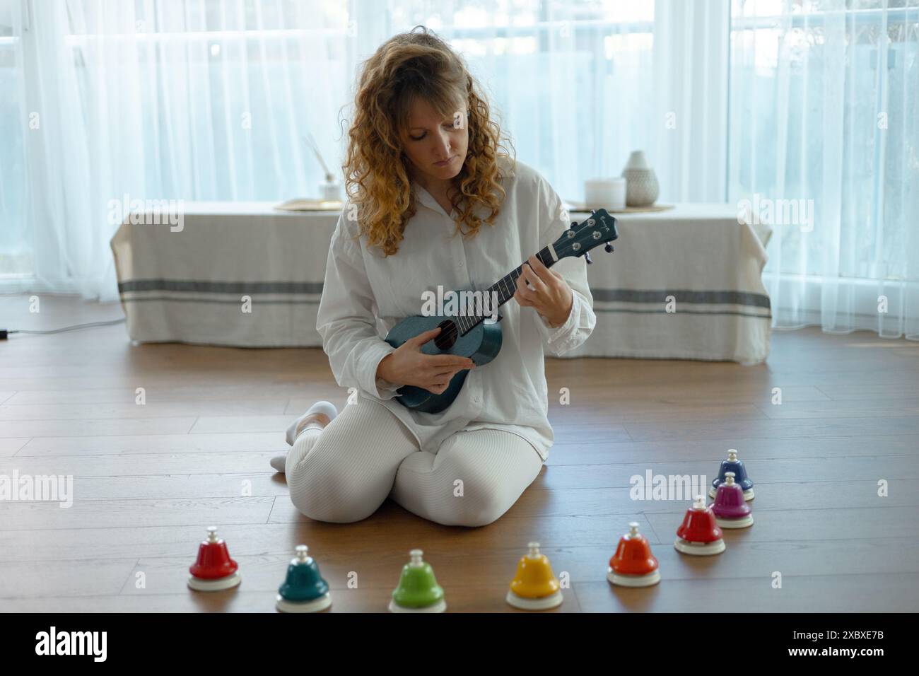 une jeune belle femme bouclée joue du ukulélé, elle est assise sur le sol dans un studio lumineux Banque D'Images