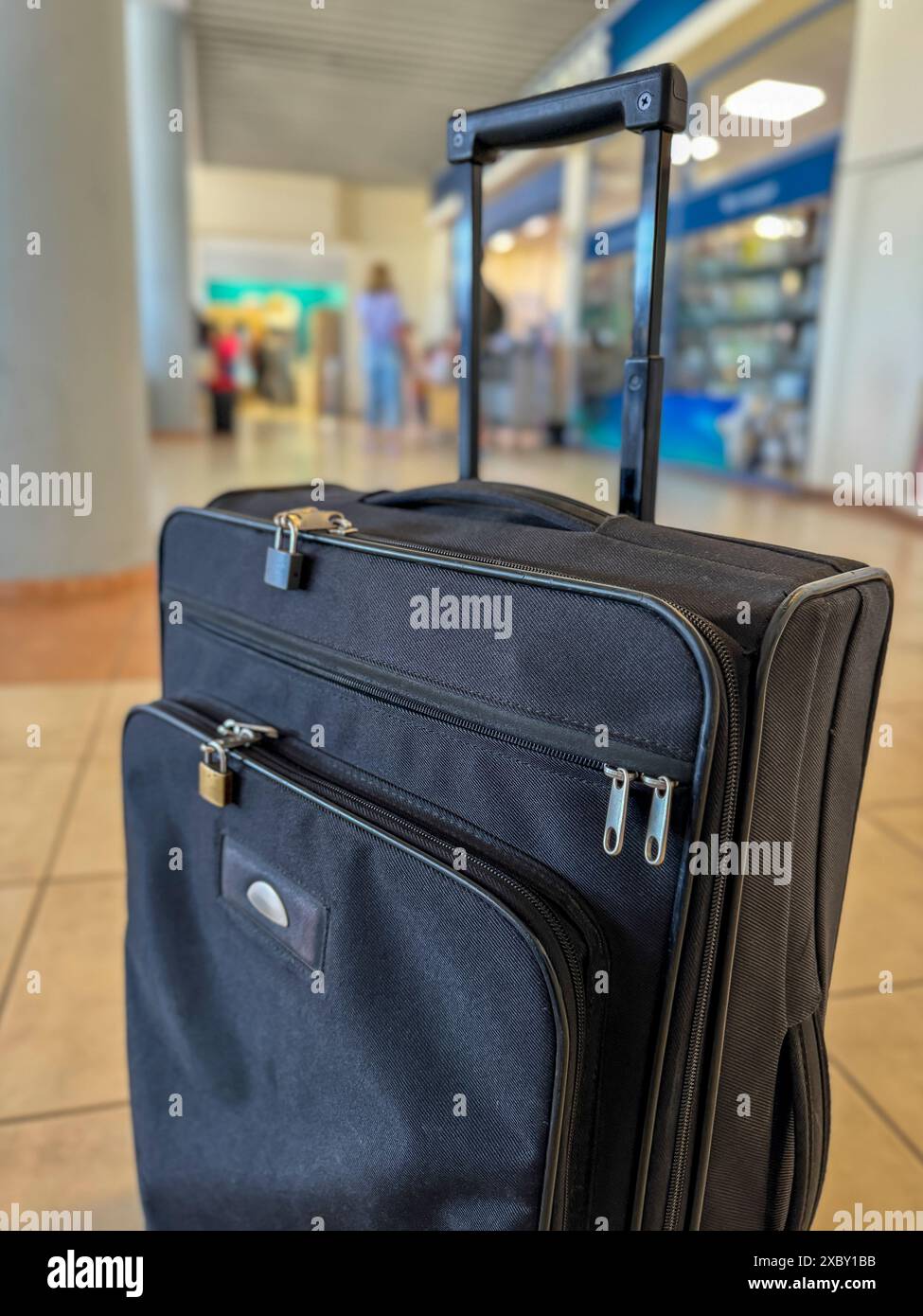 Une mallette de voyage noire et douce se dresse au premier plan, avec une boutique hors taxes floue de l'aéroport en arrière-plan, soulignant le début du voyage. Banque D'Images