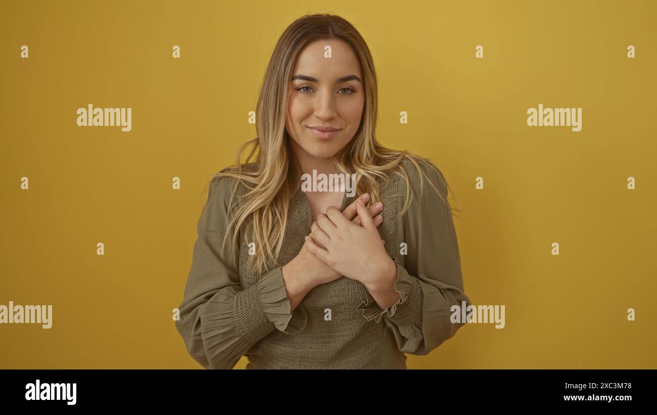 Belle jeune femme blonde dans une tenue vert olive avec ses mains sur sa poitrine, debout isolée sur un fond jaune. Banque D'Images