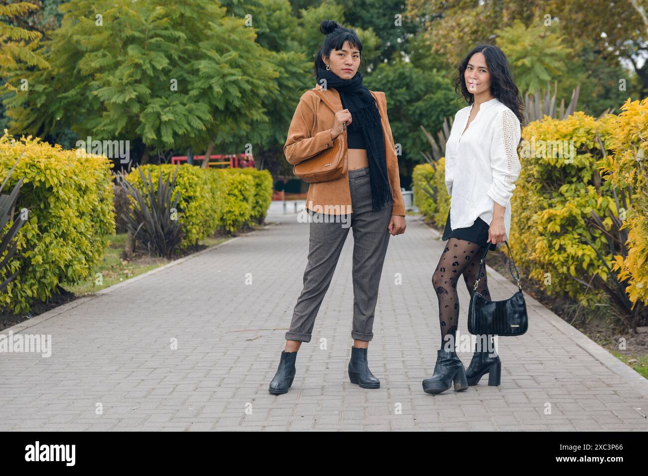 Deux élégantes jeunes femmes latino-américaines debout dans le parc posant en regardant la caméra. L'un d'eux porte une veste marron et un pantalon noir, le o Banque D'Images