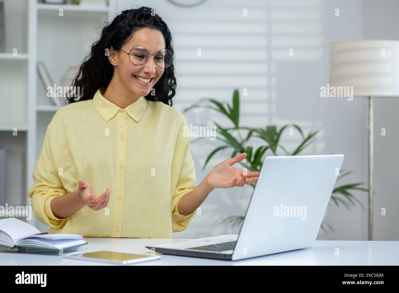 Jeune femme hispanique souriante assise dans un bureau lumineux moderne, étudiant et travaillant sur un ordinateur portable, parlant sur un appel vidéo, faisant des gestes avec les mains. Banque D'Images