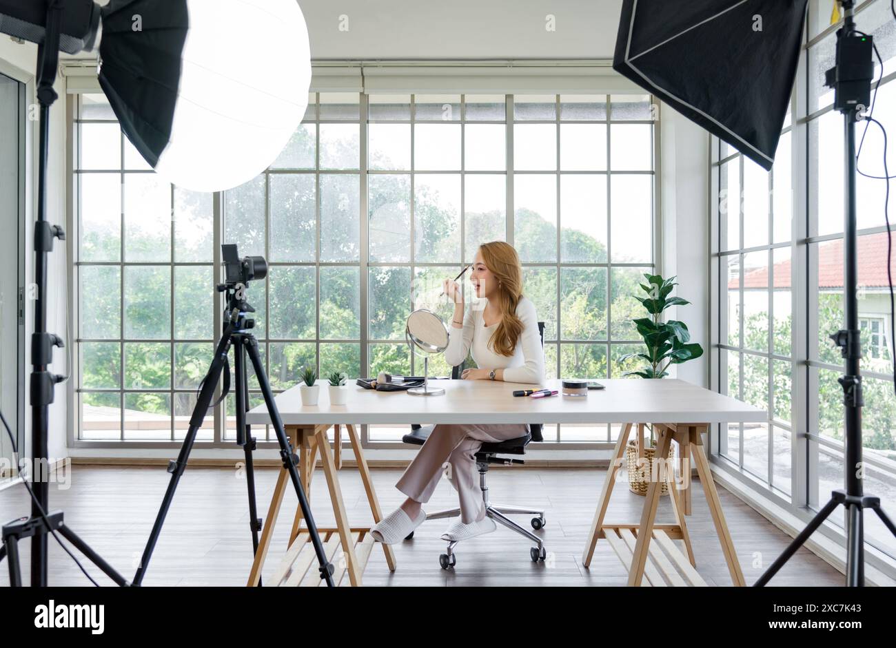 Une femme filmant son tutoriel de maquillage dans une pièce lumineuse avec une grande fenêtre, en utilisant une configuration de studio qui comprend une caméra montée sur un trépied, équipement d'éclairage Banque D'Images