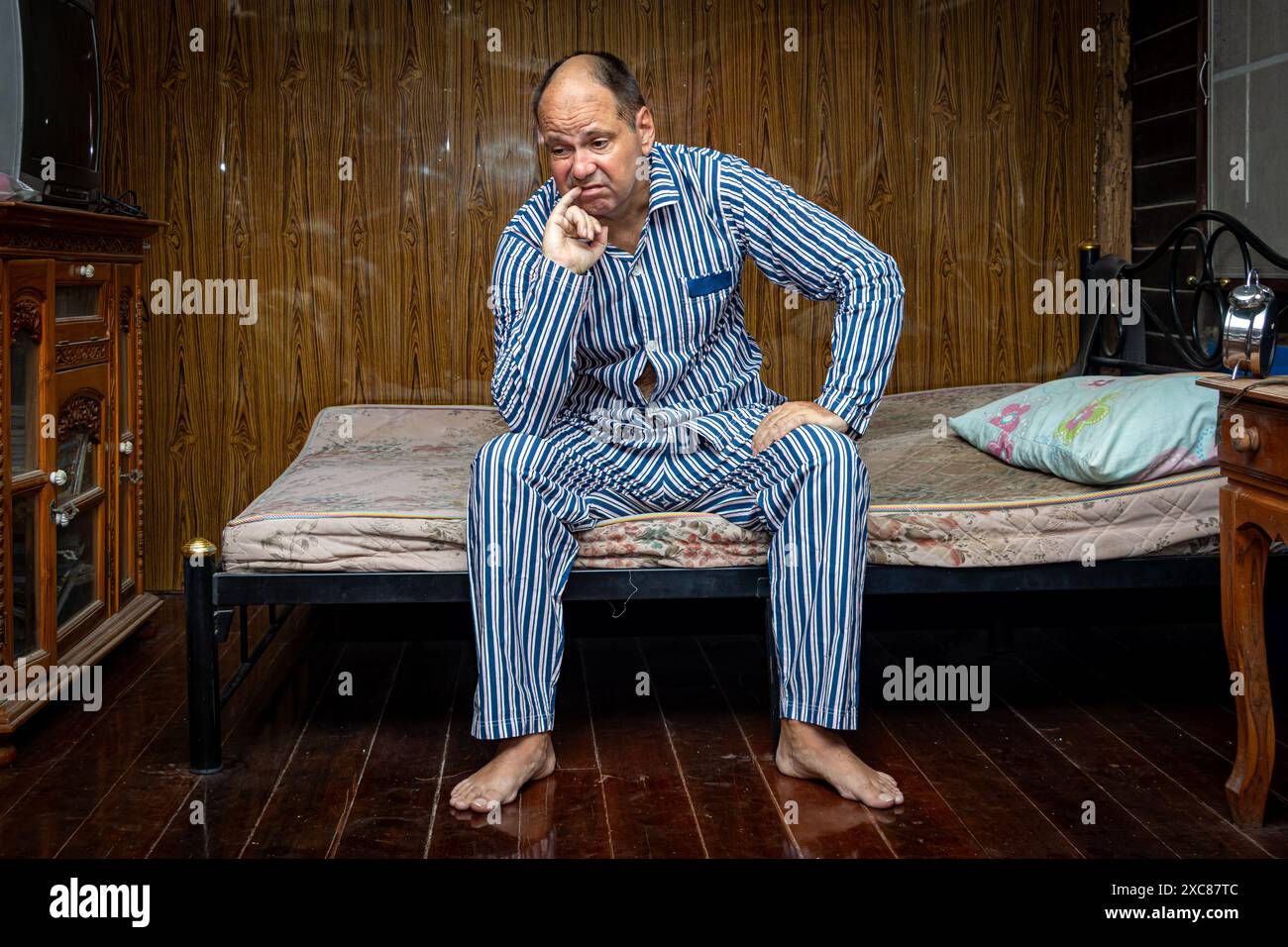 Homme grincheux en pyjama assis sur un lit de matelas dans une chambre avec vieux meubles poussiéreux Banque D'Images