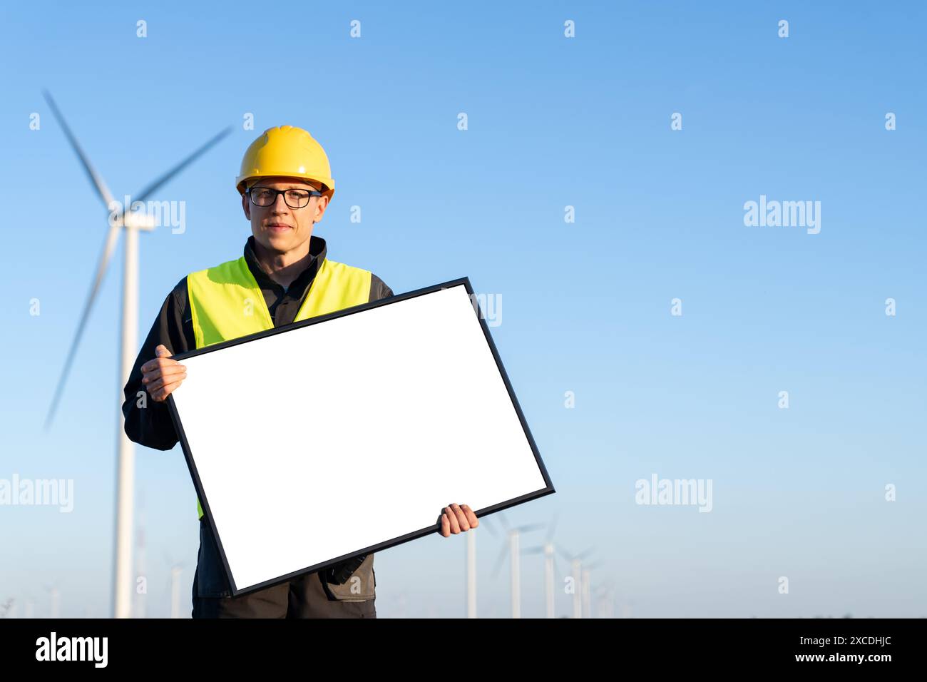 Travailleur en casque avec un tableau blanc vierge sur un champ d'éoliennes. Espace vide, vous pouvez ajouter votre texte ici. Banque D'Images