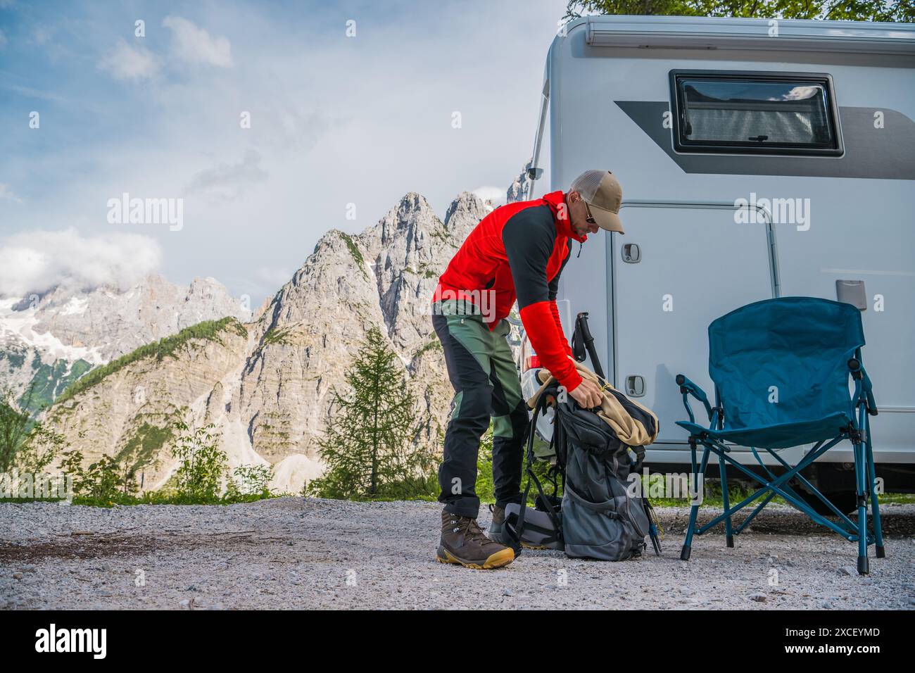 Un homme emballant son sac à dos près d'un camping-car. Il est debout sur une route de gravier devant une grande chaîne de montagnes. Il se prépare à partir en randonnée Banque D'Images