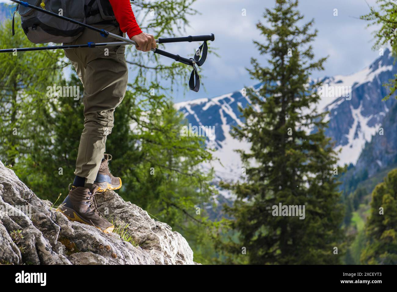 Un randonneur portant une chemise rouge et portant un sac à dos avec des bâtons de trekking marche sur un sentier rocheux avec une chaîne de montagnes enneigée en arrière-plan. Banque D'Images
