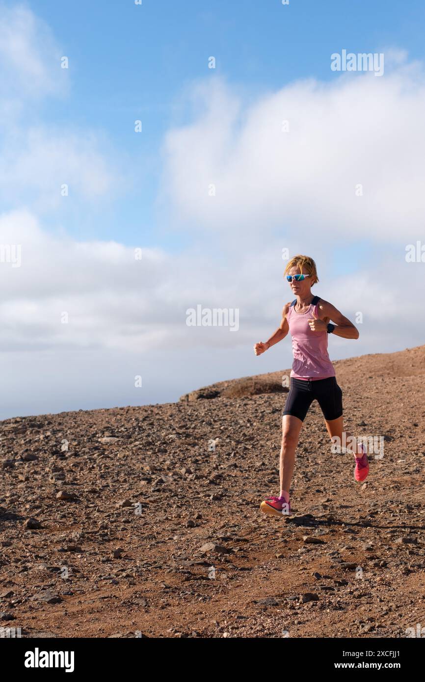 Un coureur dévoué dans un débardeur rose et un short noir navigue sur un chemin rocheux rugueux sous un ciel partiellement nuageux. Banque D'Images