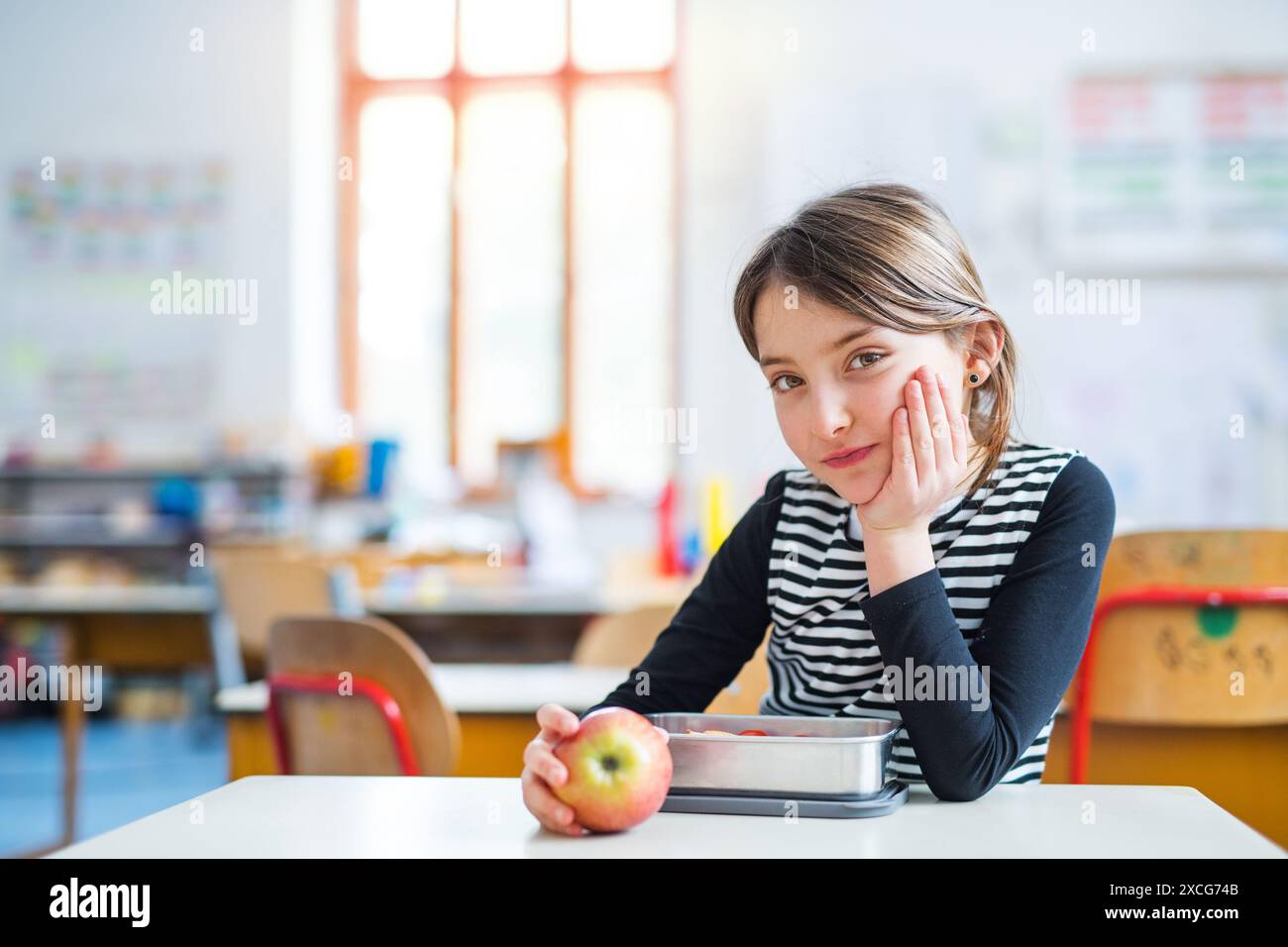 Portrait d'écolière, élève assis au bureau dans une salle de classe vide à l'école primaire. Étudiante tenant une pomme pendant l'heure de la collation, pause. Banque D'Images