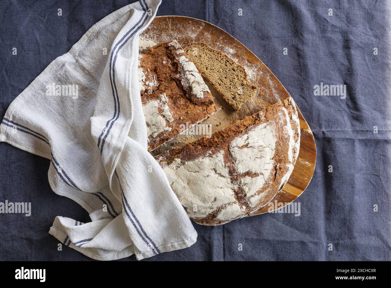 Photographie de table d'un pain de seigle frais cuit coupant en deux et tranchant sur une planche à découper en bois d'acacia recouverte d'une serviette de cuisine blanche sur un b foncé Banque D'Images
