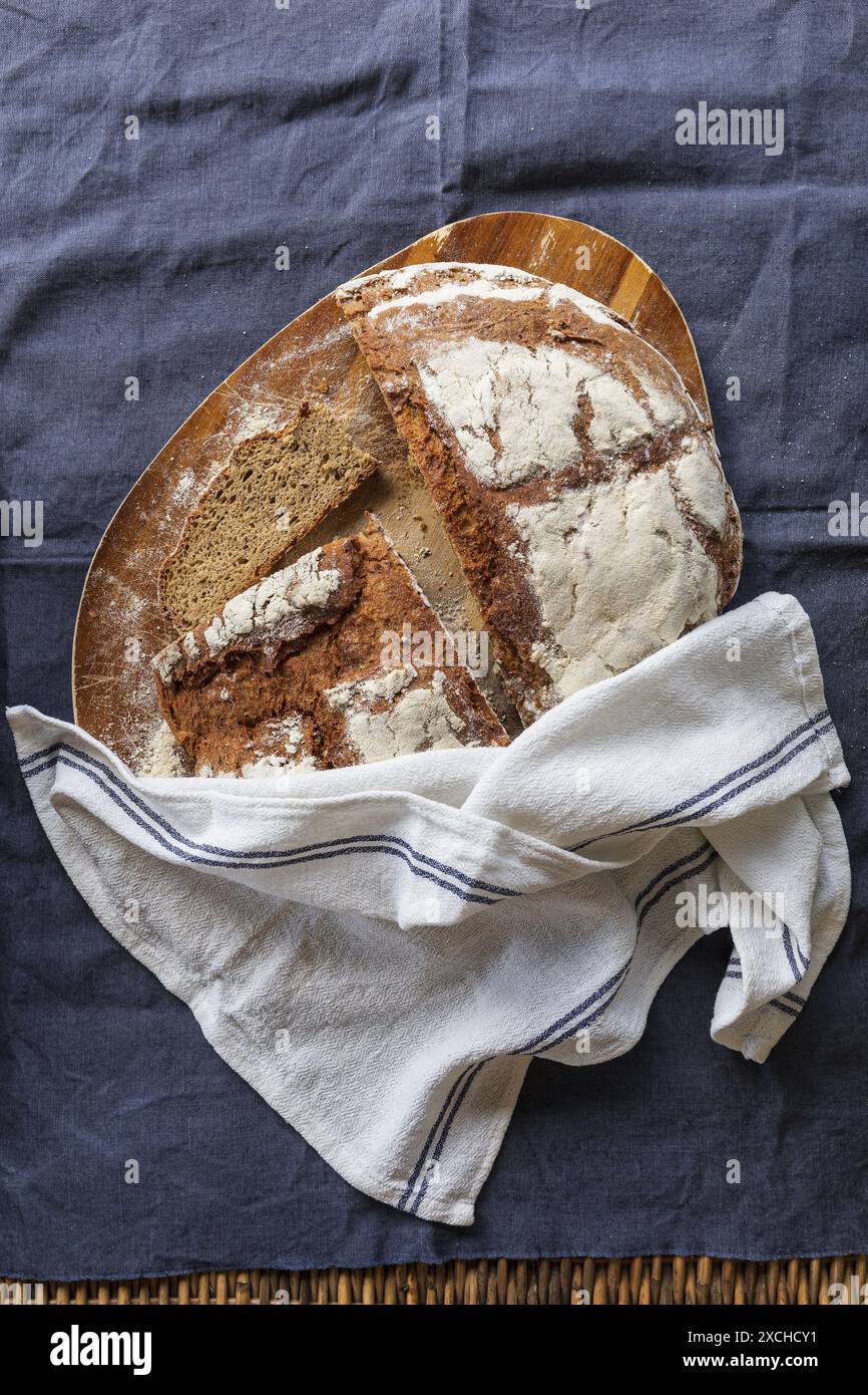 Photographie de table d'un pain de seigle frais cuit coupant en deux et tranchant sur une planche à découper en bois d'acacia recouverte d'une serviette de cuisine blanche sur un b foncé Banque D'Images